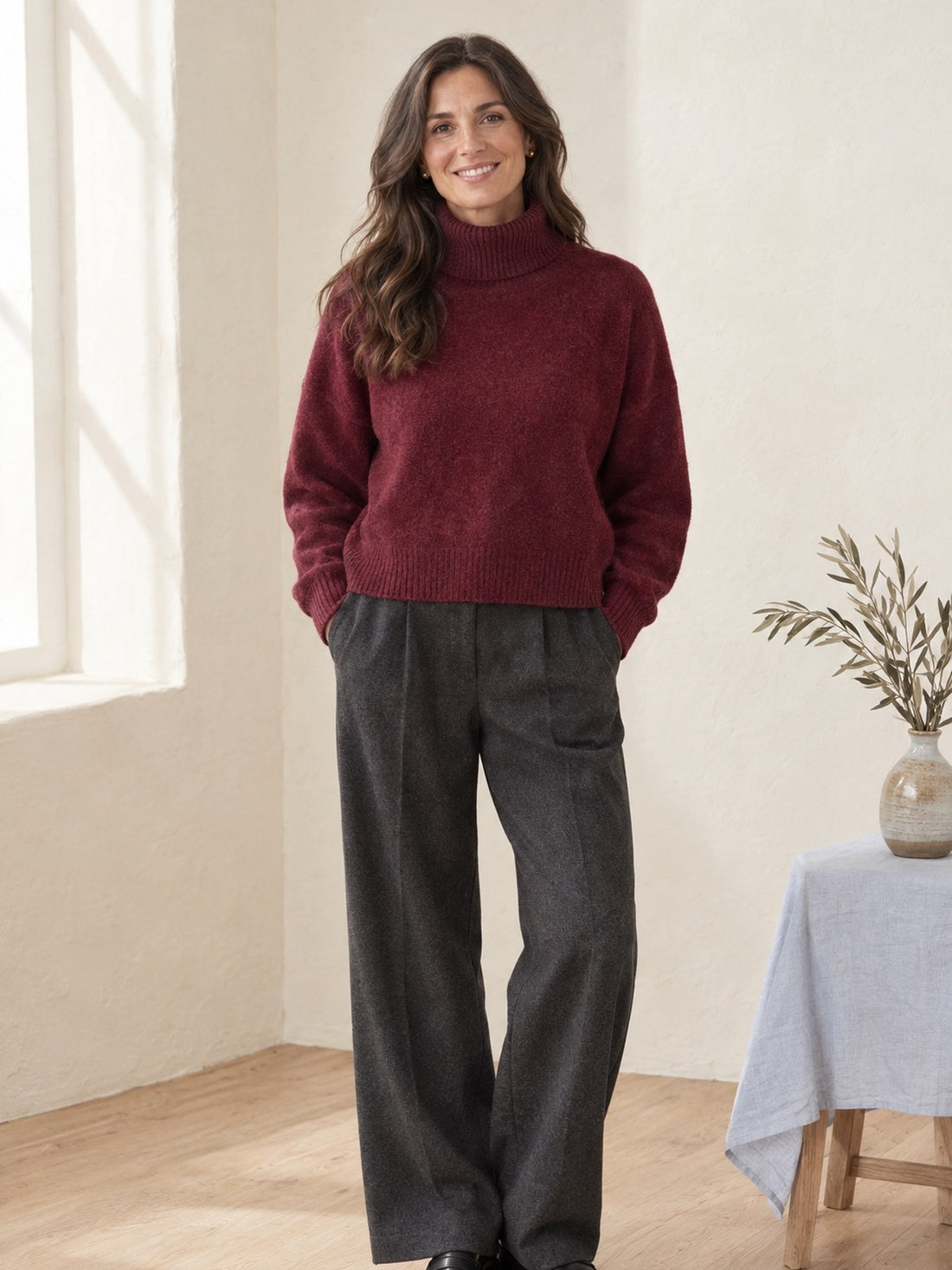 Woman in her forties wearing the burgundy Relaxed Turtleneck Jumper with grey trousers
